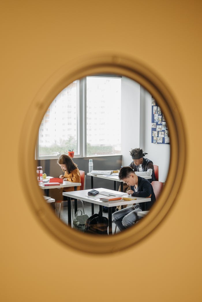 Three children concentrating on assignments in a classroom viewed through a circular window.